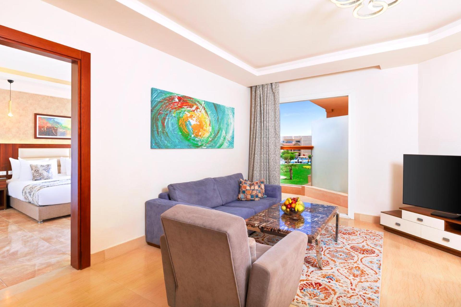 Hotel Pickalbatros Royal Moderna Sharm "Aqua Park" Esterno foto The photo depicts a modern and well-lit living room. It features a comfortable sofa in a blue tone, accompanied by an armchair in a lighter shade. A glass coffee table sits in the center, topped with a bowl of fruit. The walls are painted in a light 
