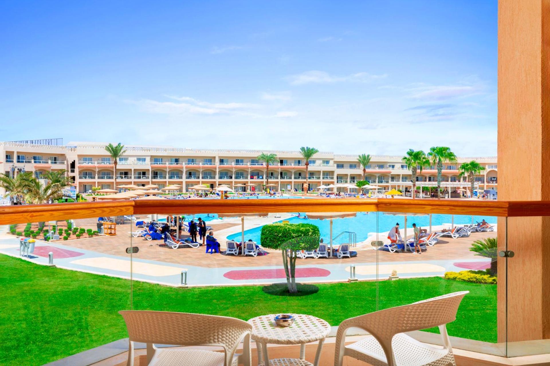 Hotel Pickalbatros Royal Moderna Sharm "Aqua Park" Esterno foto The photo shows a beautiful view from a balcony overlooking a pool area at a resort. In the foreground, there are two white chairs and a small table on the balcony. Below, the pool is surrounded by sun loungers and umbrellas, and there are several pe
