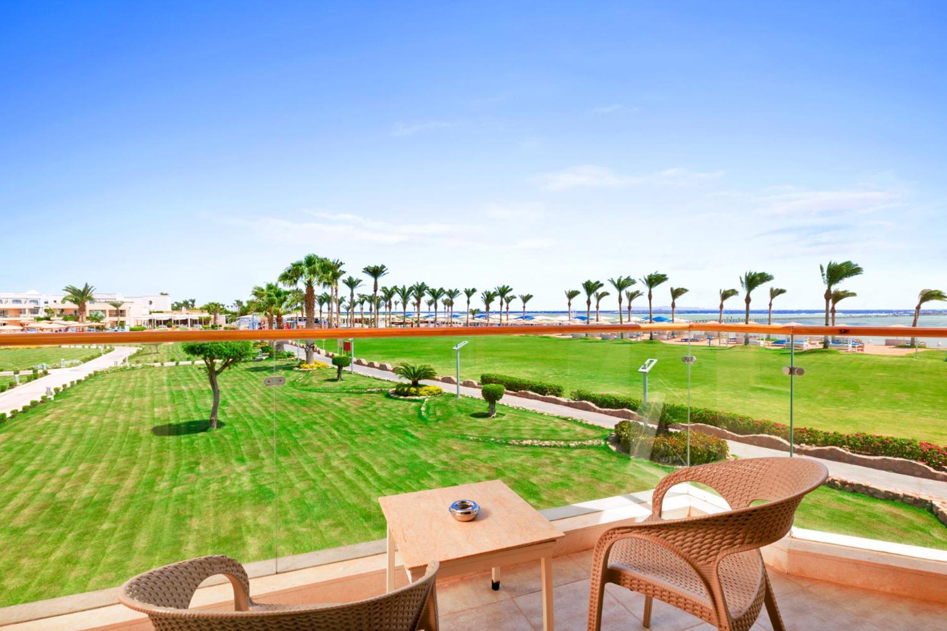 Hotel Pickalbatros Royal Moderna Sharm "Aqua Park" Esterno foto The photo shows a scenic view from a balcony overlooking a lush green lawn. There are two outdoor chairs and a small table on the balcony. In the background, palm trees line the pathway leading to the beach, which is visible in the distance under a c