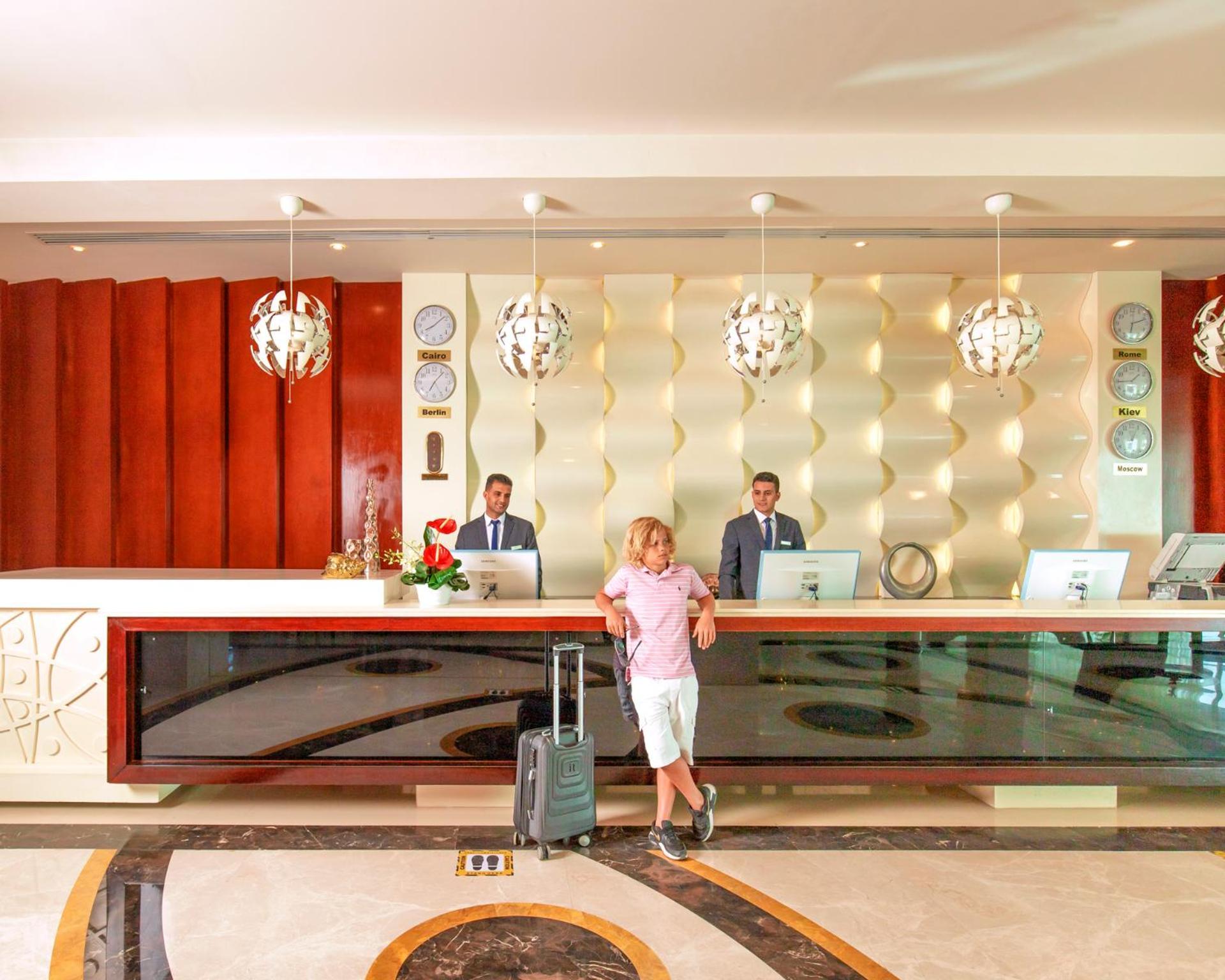 Hotel Pickalbatros Royal Moderna Sharm "Aqua Park" Esterno foto The photo depicts a hotel reception area. At the front desk, two staff members in suits are standing, presumably ready to assist guests. A man in casual attire, holding a suitcase, is standing in front of the desk. The reception has a modern design w