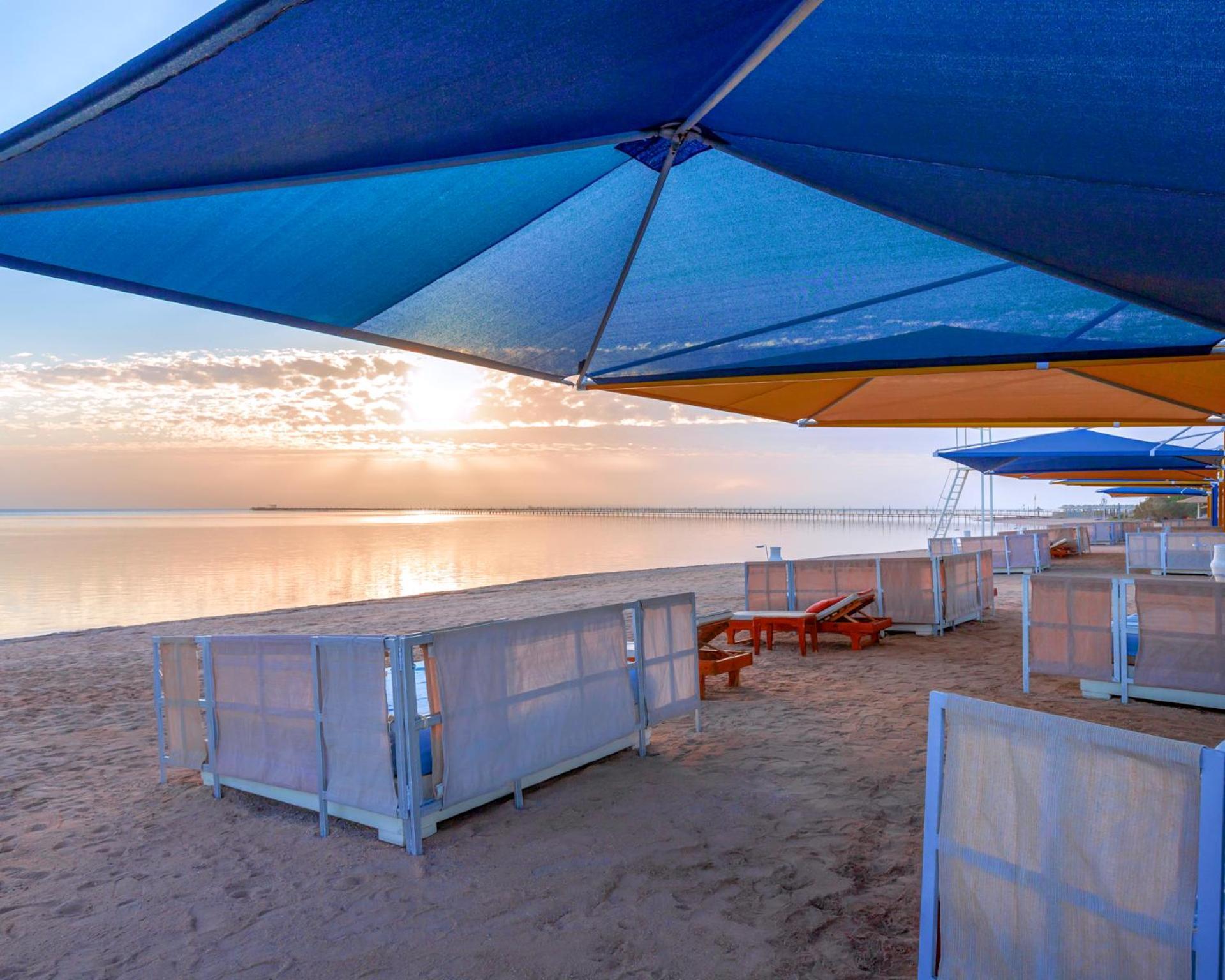 Hotel Pickalbatros Royal Moderna Sharm "Aqua Park" Esterno foto The photo shows a beach scene during sunset. There are colorful umbrellas in various shades of blue and orange set up over sandy areas. In the foreground, there are some wooden lounge chairs, and behind them, partition-like structures or beach cabana