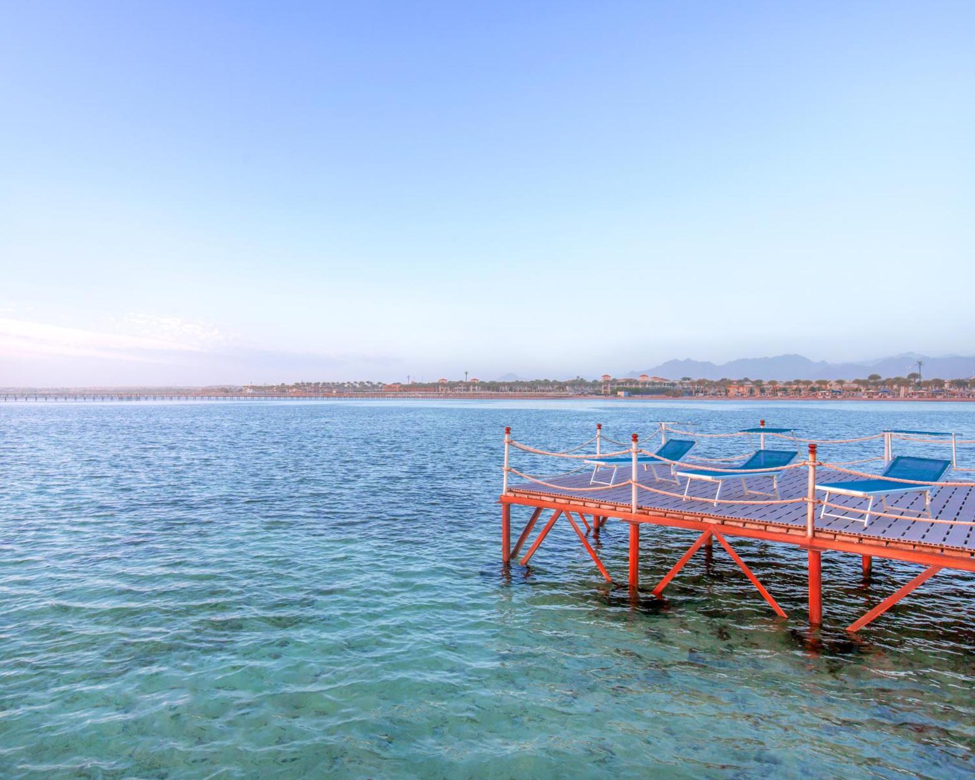 Hotel Pickalbatros Royal Moderna Sharm "Aqua Park" Esterno foto The photo shows a peaceful scene featuring a wooden dock extending into calm water. The dock is painted orange and has several blue lounge chairs placed on it, suggesting a relaxed atmosphere. In the background, there's a shoreline with some greenery