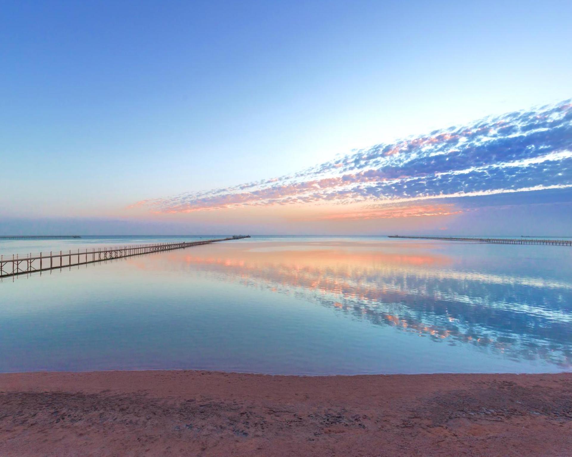 Hotel Pickalbatros Royal Moderna Sharm "Aqua Park" Esterno foto The photo depicts a serene coastal scene at dawn or dusk. There is a calm body of water that reflects the soft hues of the sky, which include shades of blue, pink, and orange. A long wooden pier extends into the water, adding depth to the composition