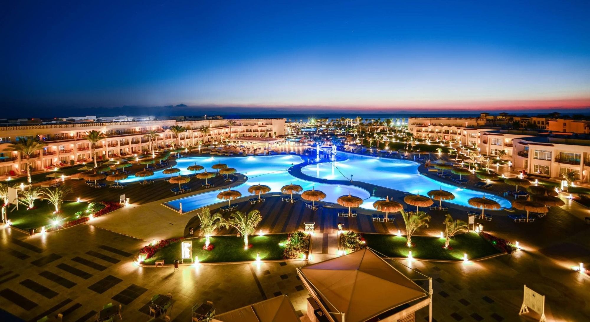 Hotel Pickalbatros Royal Moderna Sharm "Aqua Park" Esterno foto The photo showcases an elegant resort area at dusk. It features a large swimming pool, which is beautifully illuminated with blue lights, creating a serene and inviting atmosphere. Surrounding the pool are palm trees and umbrellas, enhancing the trop